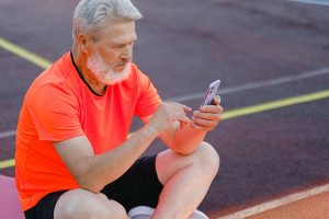older-man-in-orange-t-shirt-and-sports-look-sitting-on-the-field-with-a-smartphone